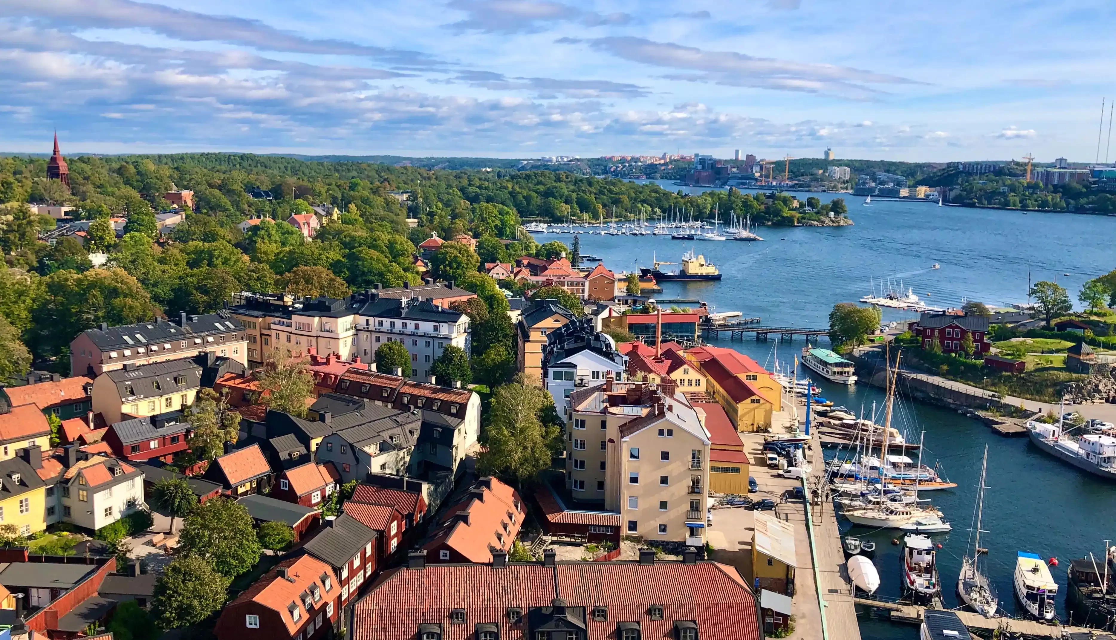 Aerial view of a coastal town with colorful buildings, surrounded by lush greenery. Boats are docked in a marina, and a body of water stretches into the distance under a partly cloudy sky.