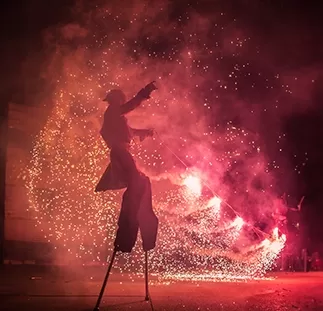 A man in costume on stilts against a smoky backdrop and fire and fireworks in the distance at a spectacle show.