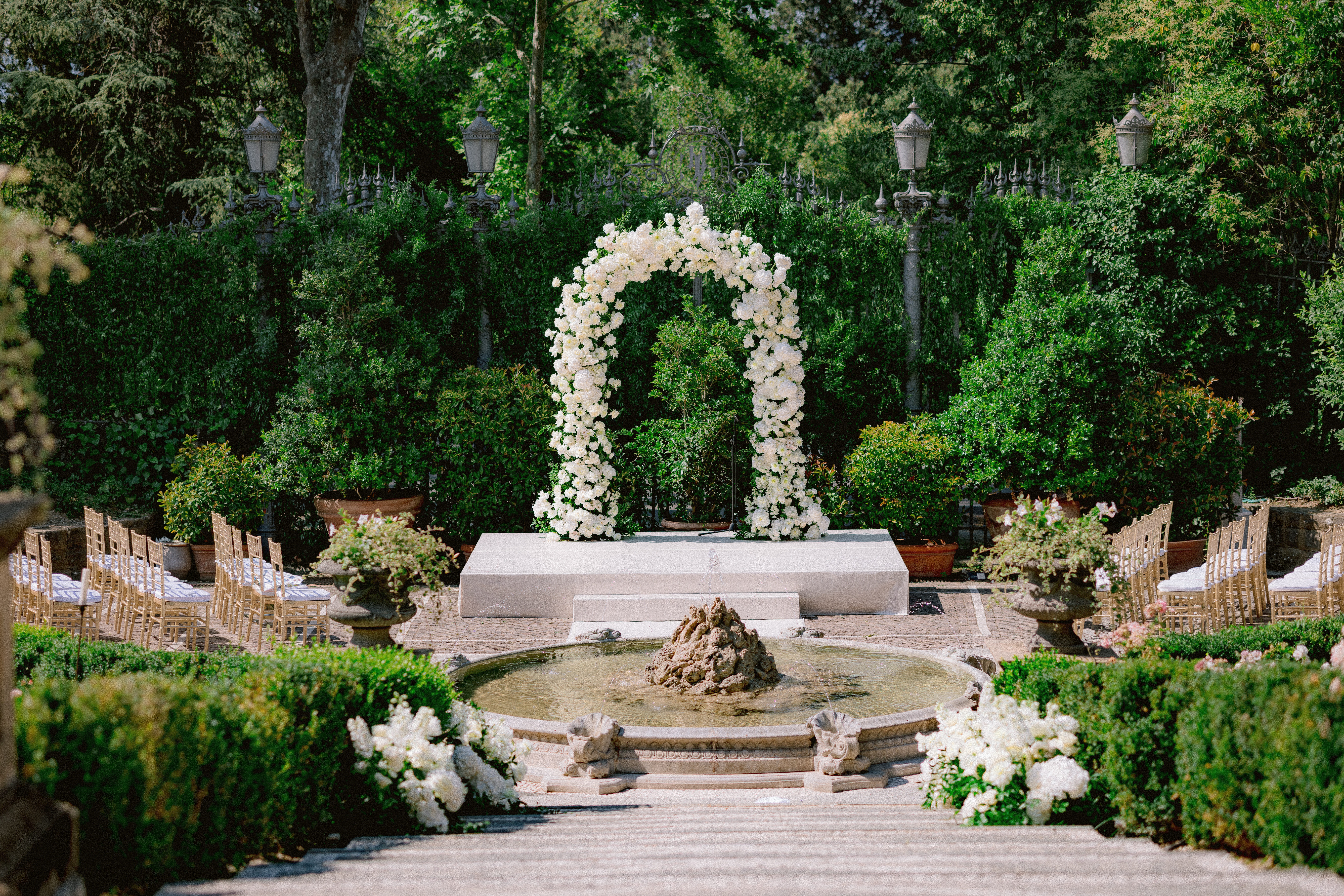 A wedding arch on a podium covered in white flowers behind a eater fountain and surrounded by chairs for wedding guests