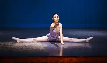 A dancer in a purple leotard performs a split on stage, extending one arm forward. The stage background is blue.