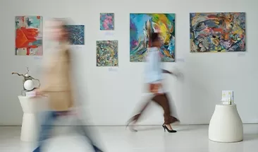 A lively bright gallery with colourful pictures on the wall and people walking past.
