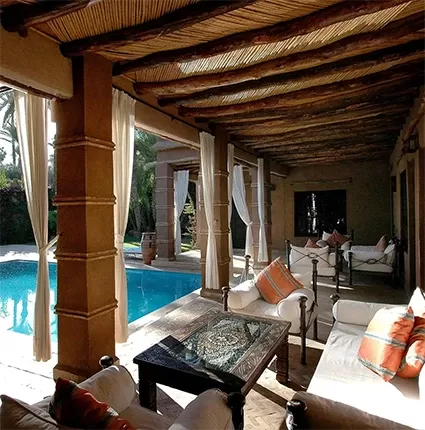 A luxurious outdoor patio with wooden beams and white drapes overlooks a shimmering blue pool. The space is elegantly furnished with white cushioned sofas adorned with orange pillows and a large glass-top coffee table.