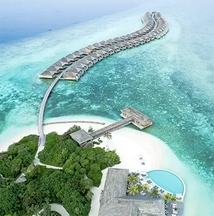 Aerial view of a tropical island resort featuring overwater bungalows extending into turquoise water, connected by a walkway. Lush greenery covers part of the island, with a swimming pool and sandy beach visible.