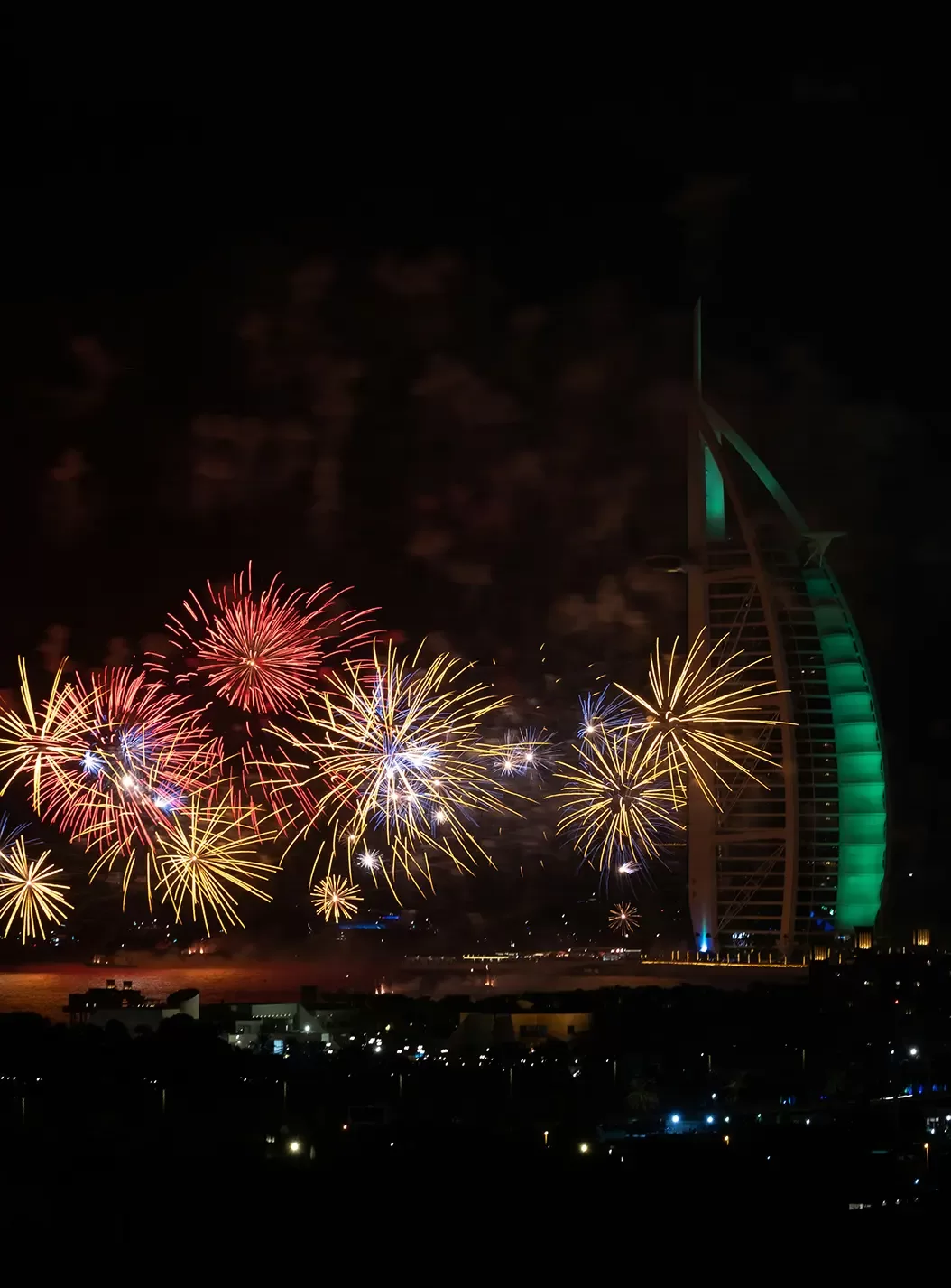 Fireworks explode in various colors over the water near the illuminated Burj Al Arab hotel in Dubai at night, with city lights visible below and the sky dark in the background.