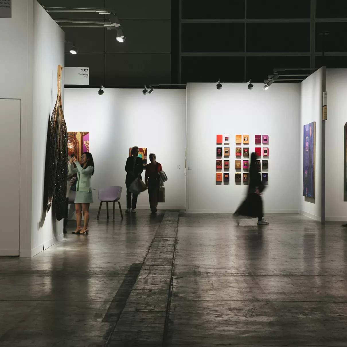 A modern art gallery with white walls displays various colorful artworks. Several people observe the exhibits, while one person walks, appearing blurred in motion. The space is brightly lit with overhead lights.