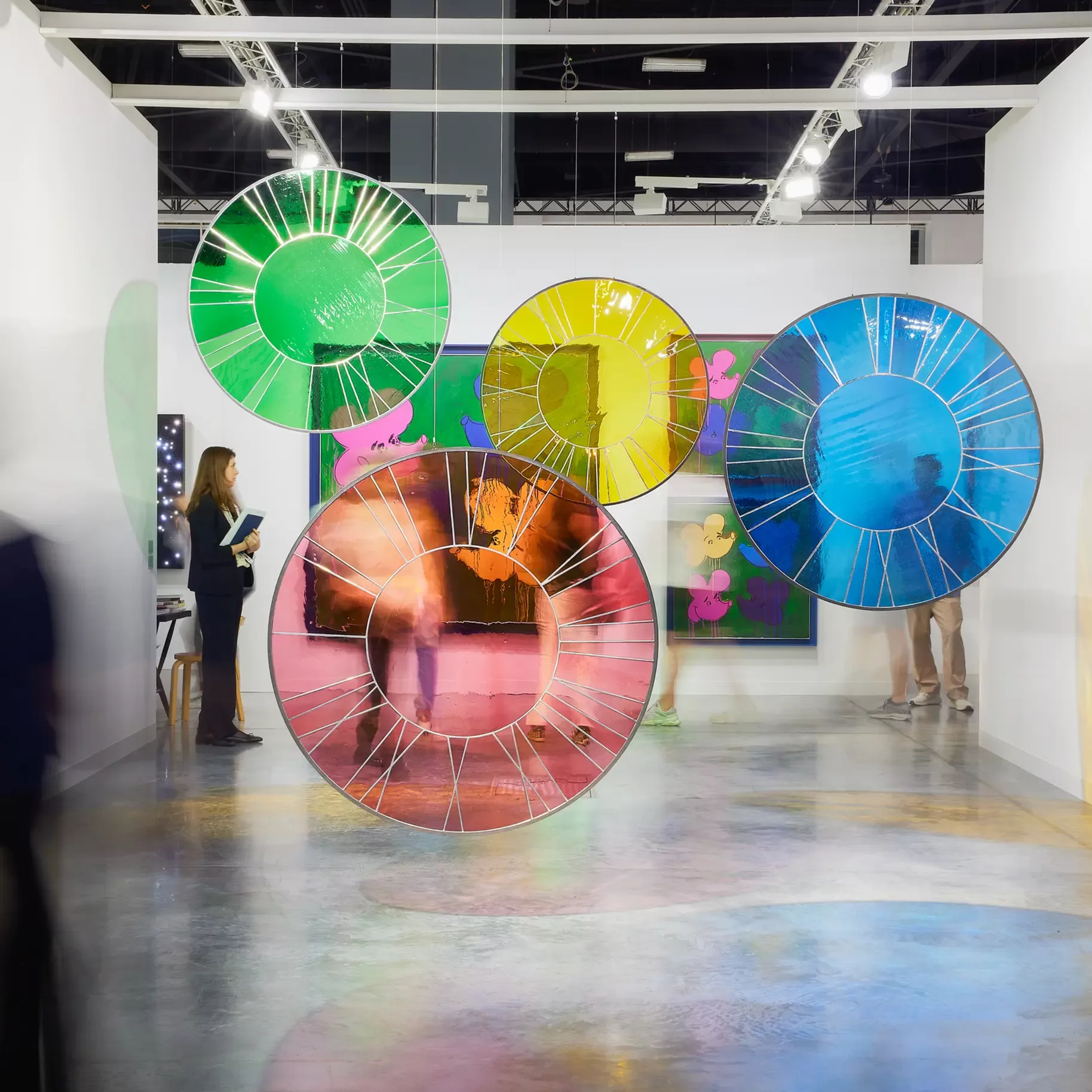 Art installation featuring transparent circular panels in green, yellow, blue, and pink. They are suspended in a gallery space with abstract paintings on the walls. A person stands to the left, viewing the exhibit. The floor reflects the vibrant colors.