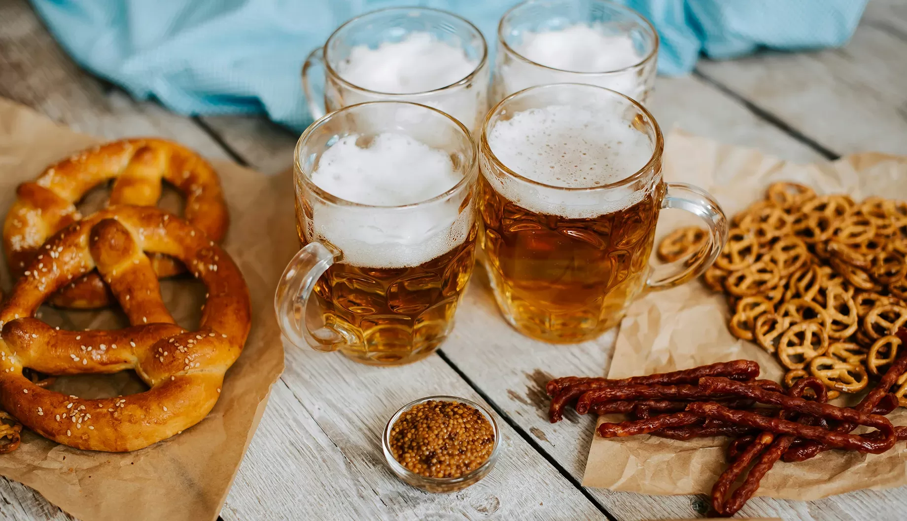 Four mugs of frothy beer are on a wooden table surrounded by large pretzels, small pretzels, mustard in a bowl, and dried sausages on parchment paper. A blue cloth is partially visible in the background.