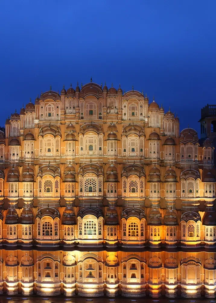 The image shows the Hawa Mahal, an iconic five-story palace in Jaipur, India, beautifully illuminated against a dark evening sky. The façade features intricate lattice windows and detailed architectural designs, creating a stunning visual effect.