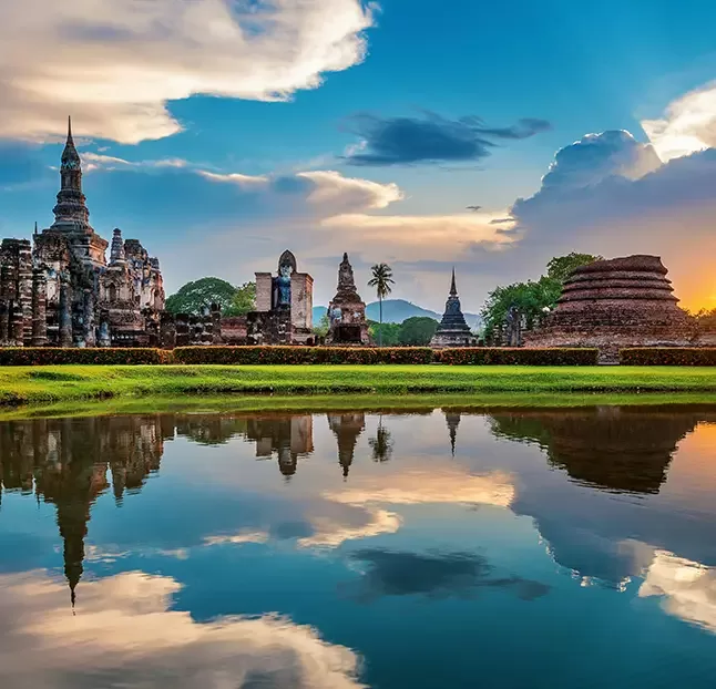 Ancient stone temple ruins at sunset with a lake in the foreground reflecting the vibrant sky, clouds, and structures. Lush greenery surrounds the scene, creating a peaceful and historical atmosphere.