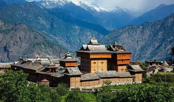 A traditional wooden temple complex nestled in a lush valley with tall, snow-capped mountains in the background. The temple features intricate architecture with multiple tiers and sloped roofs, surrounded by green foliage.