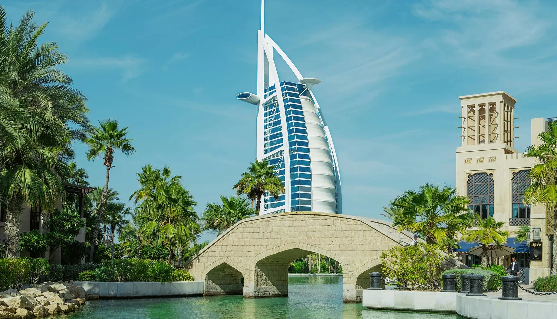 A daytime view of the Burj Al Arab hotel in Dubai, seen from a waterway lined with palm trees and traditional buildings.