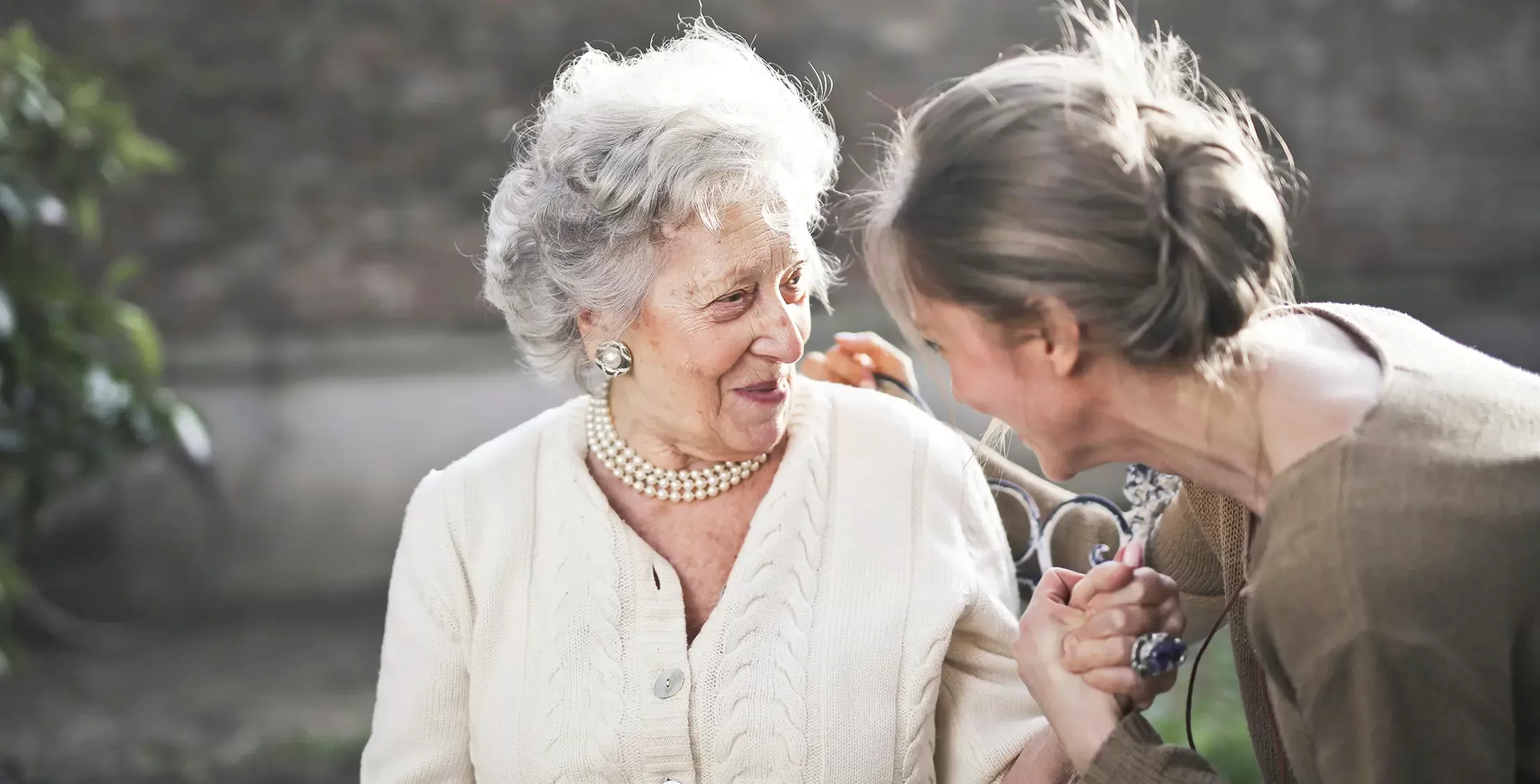 Older women wearing pearls necklace, earrings and a cable knit cardigan smiling at a younger women in the garden.