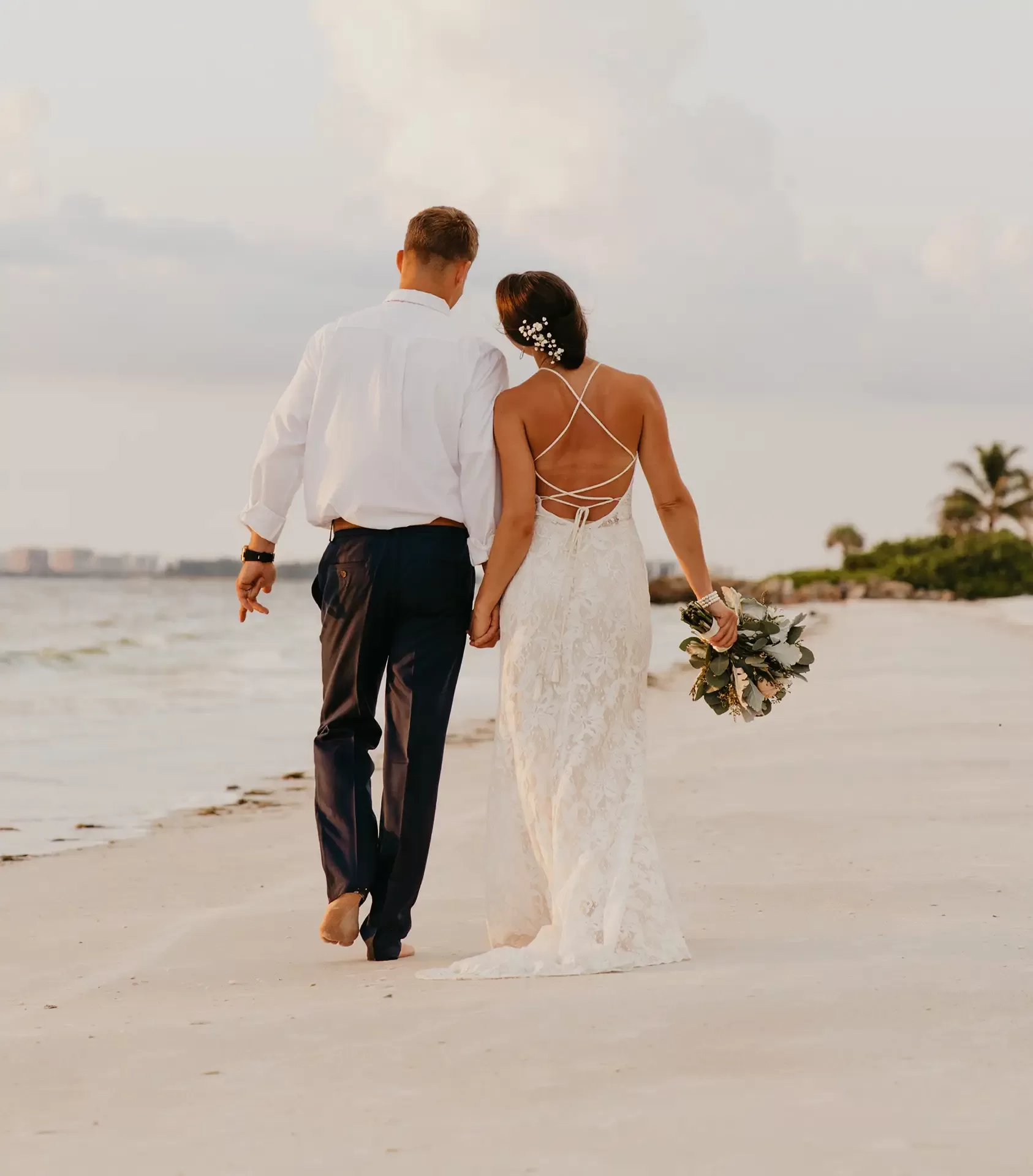 A couple walks hand in hand on a sandy beach. The bride wears a backless lace wedding dress and holds a bouquet, while the groom is in a white shirt and trousers. The ocean and sky are in the background, and a palm tree is visible to the right.