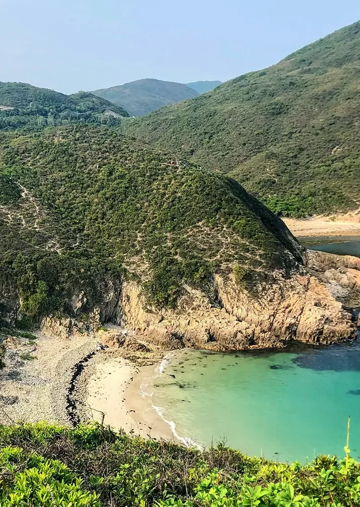 A scenic view of a secluded beach with clear turquoise water, surrounded by rocky cliffs and lush green hills under a clear blue sky. The landscape is serene and natural, with no people present.