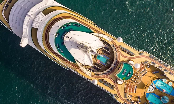 Aerial view of a large cruise ship with multiple decks featuring pools, sun loungers, and a central covered area. The ship sails on a calm, deep green sea.