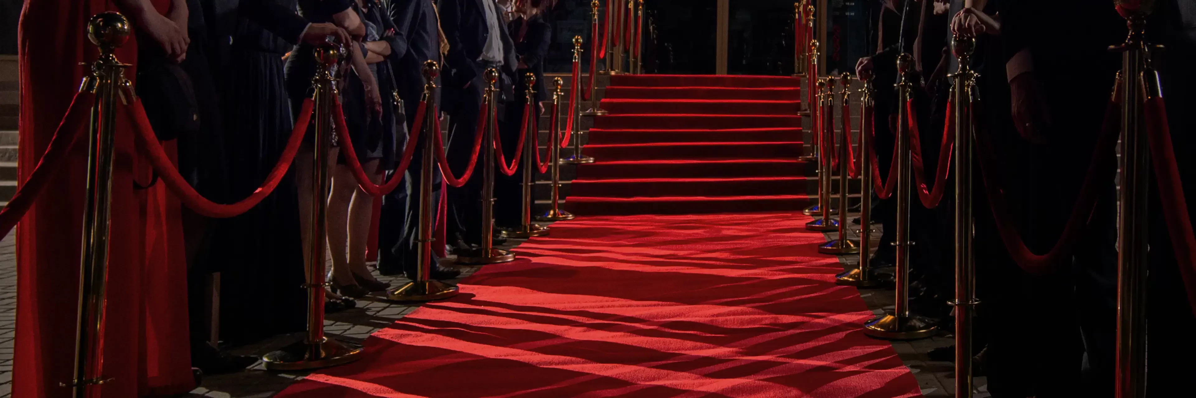 A glamorous red carpet event with a bright red carpet flanked by golden stanchions and red velvet ropes, leading up to an entrance. People in formal attire are lining the sides, creating an elegant and prestigious atmosphere.