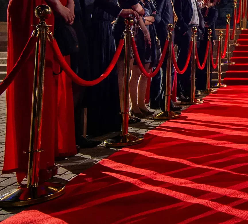 A luxurious red carpet lined with red ropes and golden stanchions leads up a flight of stairs. People in formal attire stand on either side, partially visible, creating a grand and elegant atmosphere.