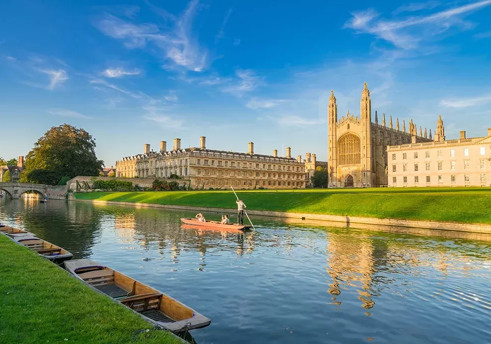A serene river scene with a person punting a boat near a historic Gothic building, likely a university or cathedral, under a clear blue sky. Green lawns and stone bridges add to the picturesque setting.