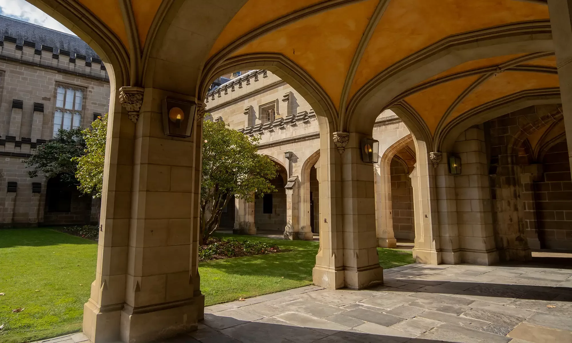 A historic stone archway leads to a courtyard with green grass and a tree. The structure features a vaulted ceiling and ornate detailing. Sunlight creates shadows on the ground, enhancing the architectural elegance of the University of Melbourne, Australia.