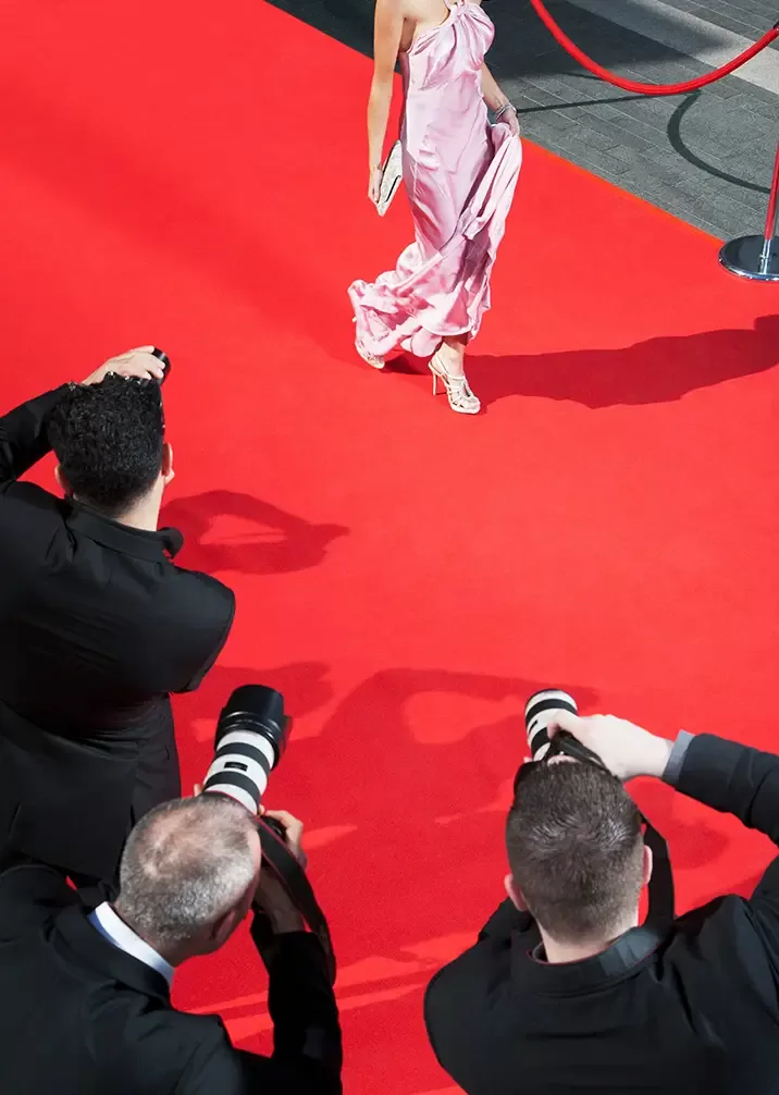 A person in a flowing pink dress walks on a red carpet, holding a clutch. Photographers in black suits with cameras gather at the edge, capturing the scene. A red velvet rope and metal stanchions line the carpet.