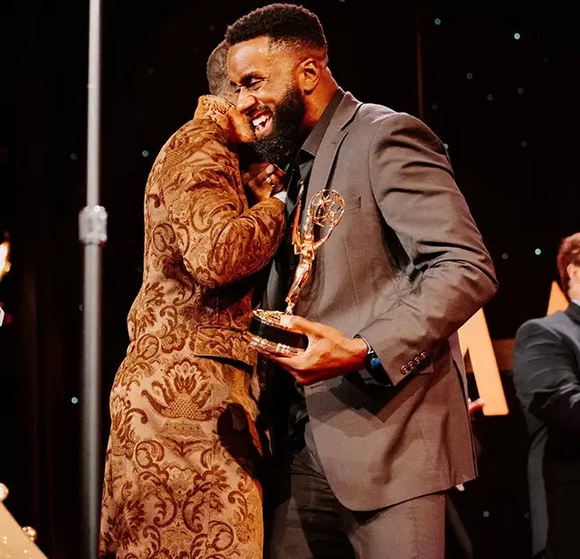 Two men stand on a stage. One wears a patterned jacket, and the other in a suit holds an award shaped like an Emmy. They are embracing, with one man smiling joyfully.