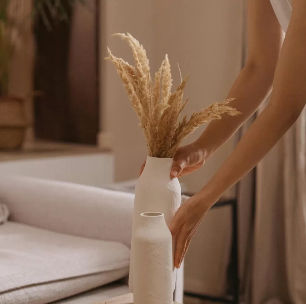 A person is placing a white ceramic vase filled with dried wheat stalks on a light-coloured surface. The background includes a blurred view of a couch and other home decor items, creating a cozy and minimalistic interior scene.