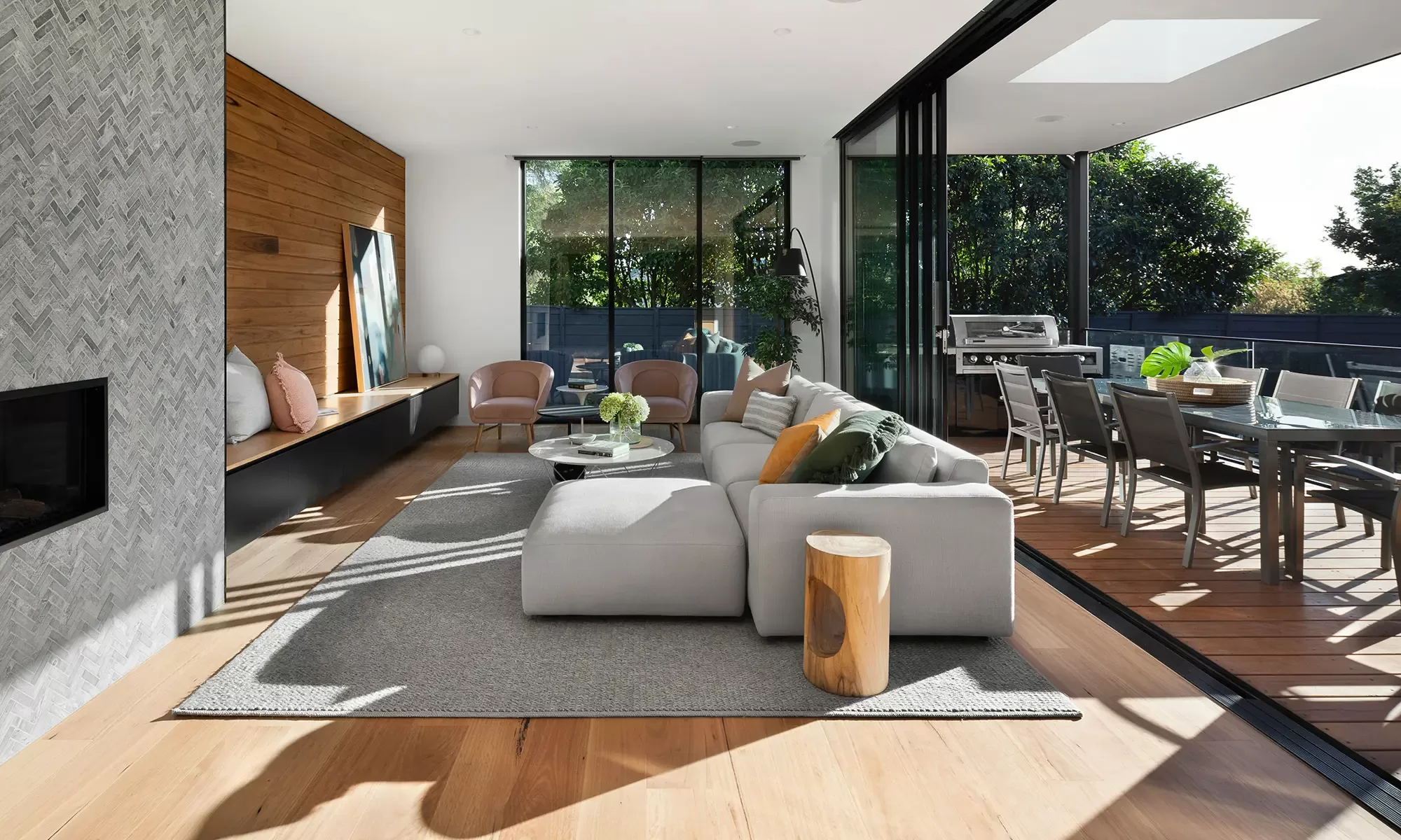 A modern living room with a sleek, light gray sofa and a wooden accent wall. Floor-to-ceiling glass doors open to a patio, which features a dining table and lush greenery. Soft natural light enhances the warm, inviting space.
