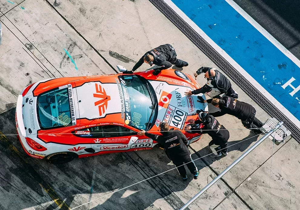 Overhead view of a racing car in a pit stop, with five crew members working around it. The car is red and white with various logos and the number 400 on its side. The scene takes place on a concrete pit lane beside a blue track boundary.