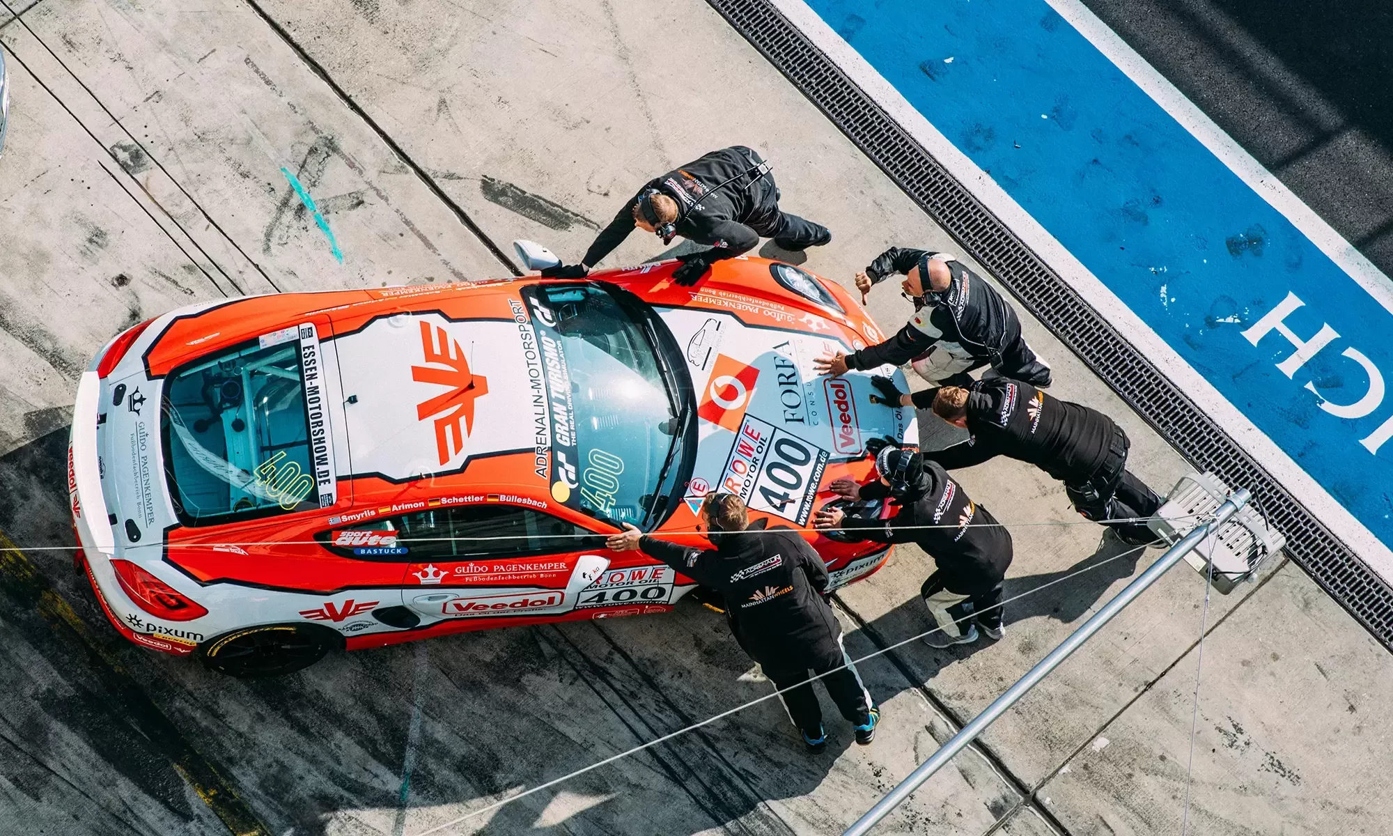 Overhead view of a racing car in a pit stop, with five crew members working around it. The car is red and white with various logos and the number 400 on its side. The scene takes place on a concrete pit lane beside a blue track boundary.