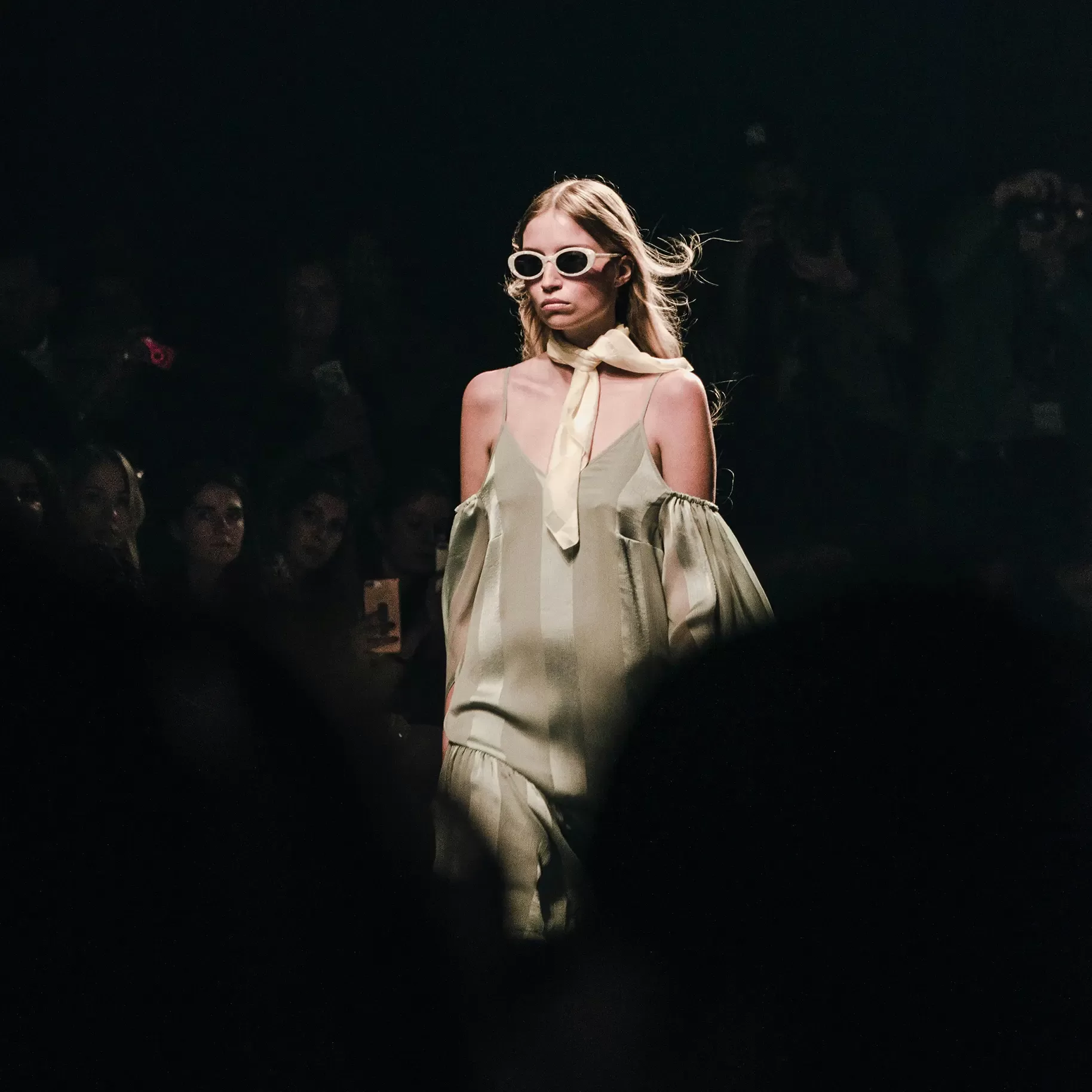 A model walks confidently on the runway wearing a pale green off-shoulder dress, a light-colored scarf, and white sunglasses. The background is dimly lit with an audience watching and capturing the event.