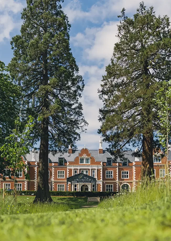 A grand, red-brick mansion with large windows and a decorative entrance is framed by two tall trees, under a blue sky with scattered clouds. The foreground features a lush green lawn.