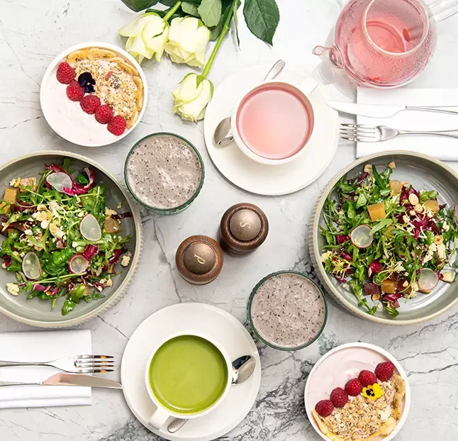 A top-down view of a meal on a marble table, including salads with greens and radishes, two smoothies, green soup, pink drinks, and a bowl of yogurt with raspberries. Nearby are white roses and neatly arranged cutlery.