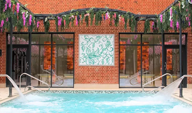 Outdoor heated pool with cascading water jets, surrounded by a brick wall adorned with hanging purple and green flowers. Large windows in the background reflect the interior lighting at The Fairmont, Windsor.