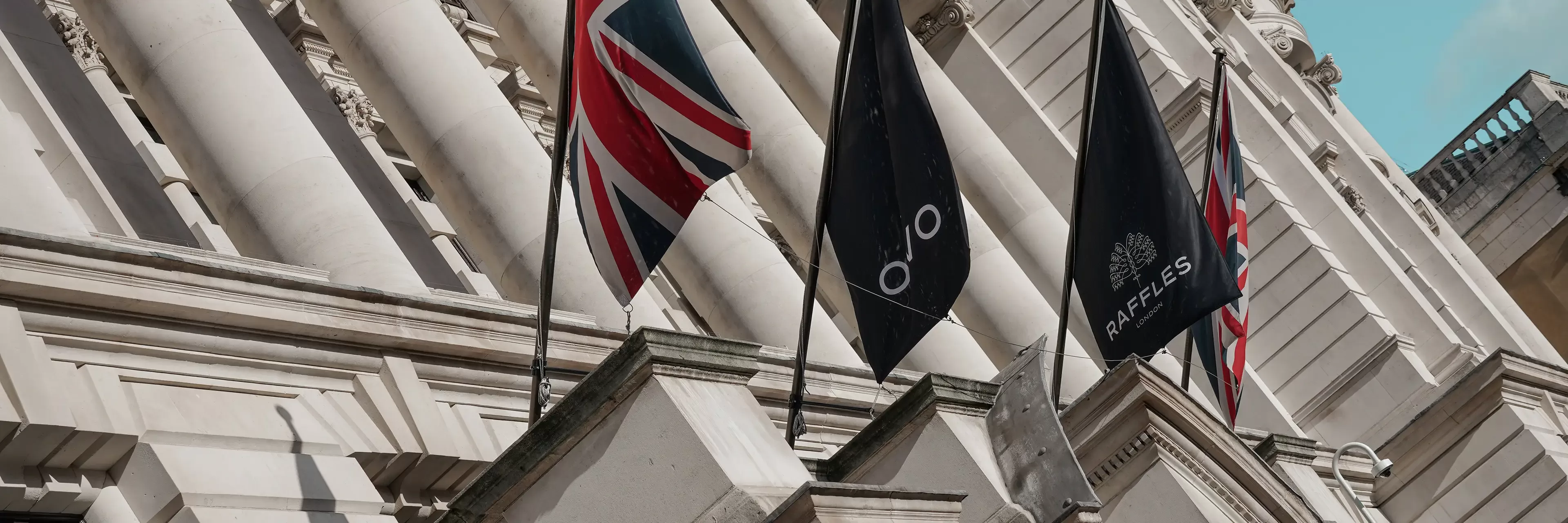 Three flags hang in front of an ornate white building with columns. The left flag is the Union Jack, and the two black flags display the Raffles hotel logo. Another Union Jack is partially visible on the right.
