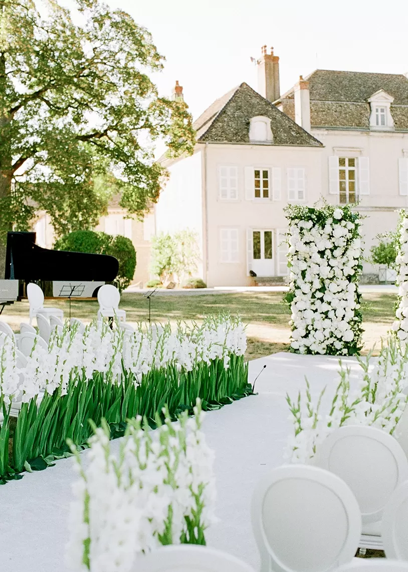 Estate garden wedding ceremony decor with white aisle, white flowers and white chairs with grand piano outside of grande estate.