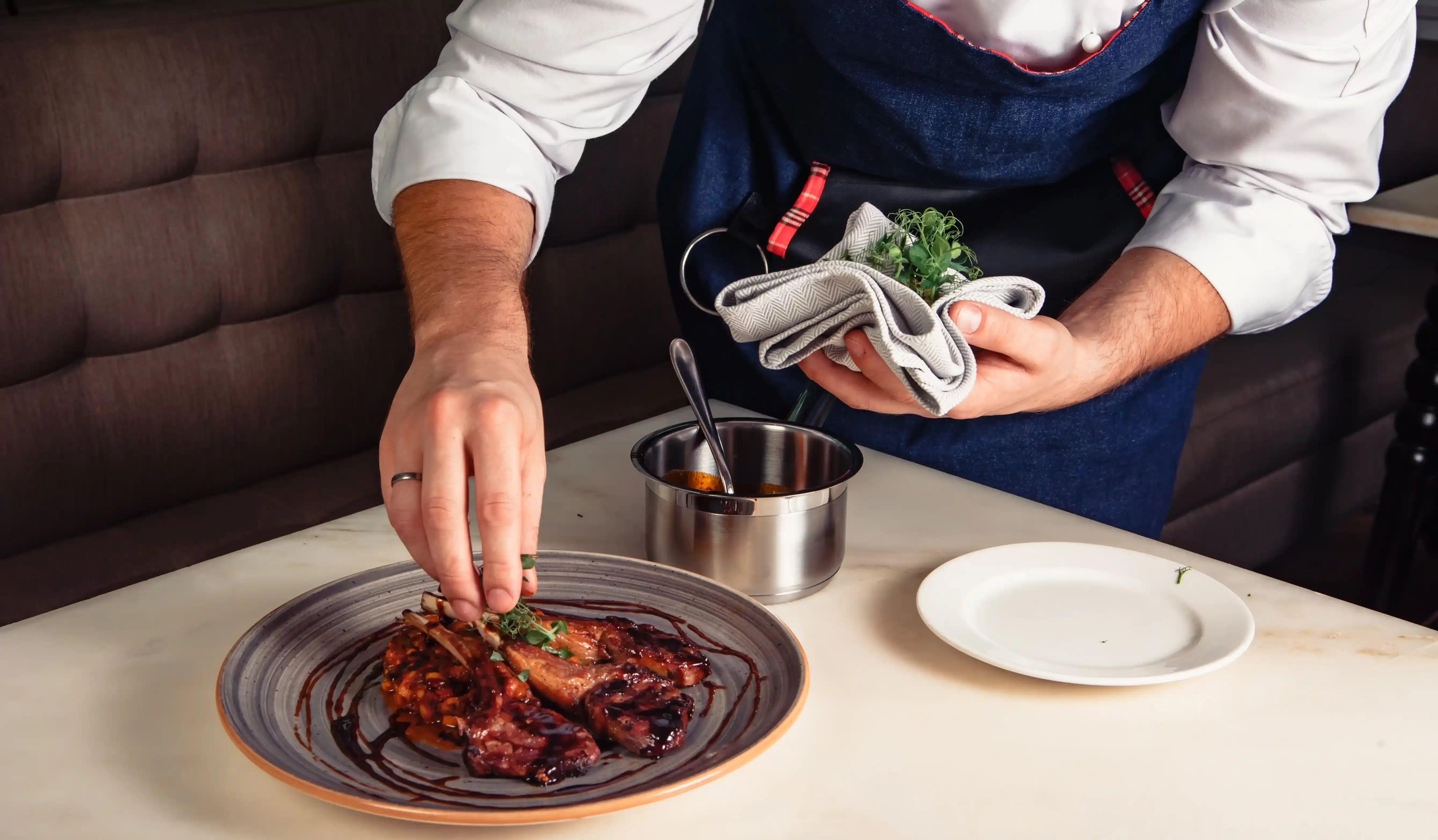 A chef in a dark apron and white shirt prepares a gourmet dish, adding garnish to a plate with meat drizzled in sauce. A small pot and empty white plate are on the table, with a plush booth seat in the background.