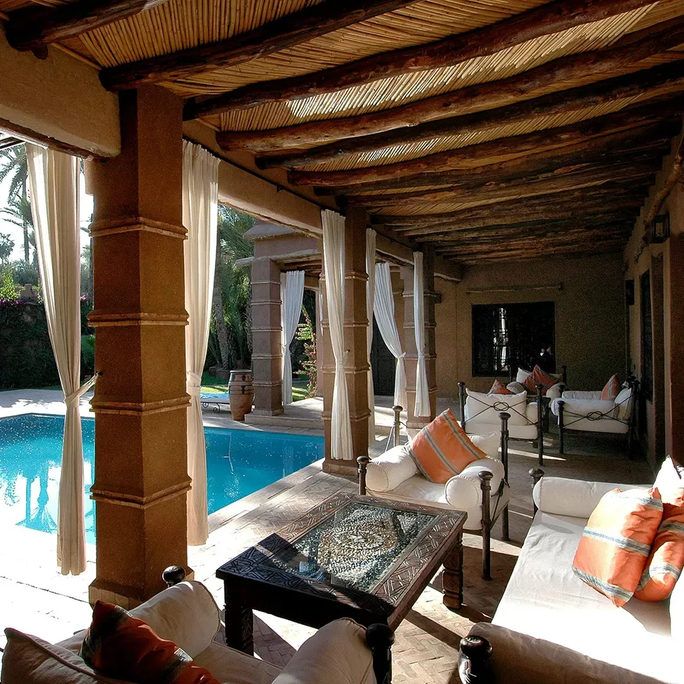 A shaded patio with sofas and a glass-top table overlooks a clear blue swimming pool, surrounded by wooden beams and curtains.