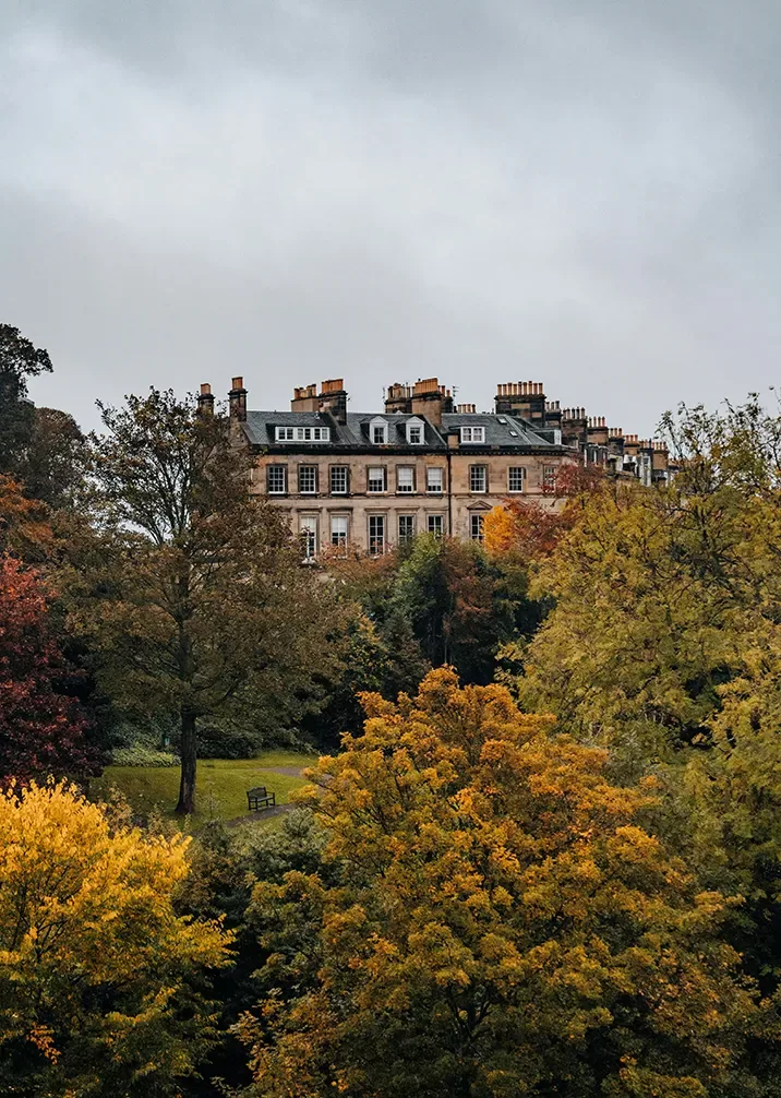 A stately, multi-story building with numerous chimneys sits among trees displaying autumn foliage in hues of green, yellow, and orange. The scene is overcast, giving the image a serene and tranquil atmosphere.