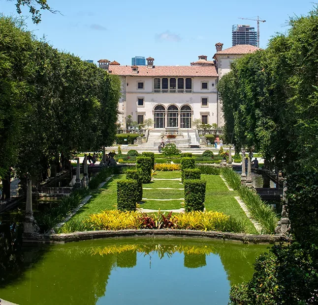 A grand estate with a large mansion in the background and meticulously manicured gardens in the foreground. The garden features symmetrical hedges, vibrant flowerbeds, and a tranquil pond reflecting the greenery. Skyscrapers are visible in the distant skyline.