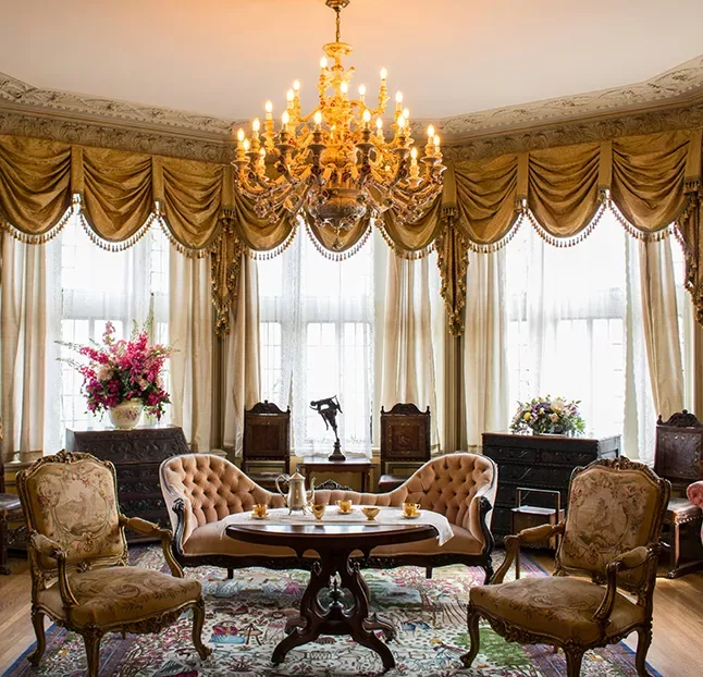 Elegant living room with ornate furniture, including four upholstered chairs around a small wooden table. A grand chandelier hangs from the ceiling. The windows are adorned with golden drapes, and vases with colourful flowers are placed around the room.