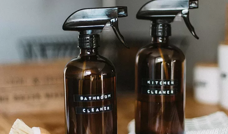 Two dark amber spray bottles with black pump tops stand side by side on a countertop. One bottle is labelled "Bathroom Cleaner" and the other "Kitchen Cleaner" in white letters on black tape. A blurred wooden box and some cleaning items are visible in the background