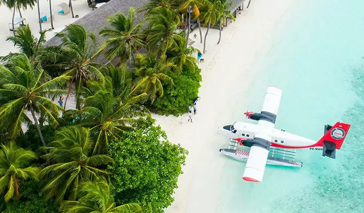 A seaplane with white and red markings docked on the turquoise waters of a tropical beach. The scene is surrounded by lush palm trees and a white sandy shore, creating a serene vacation setting in an island resort.
