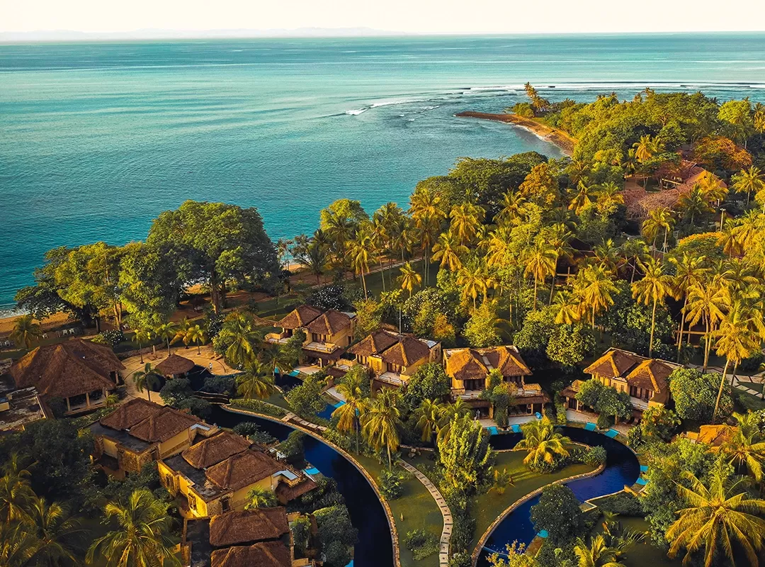 Birdseye view of luxury resort lodges amognst palm trees and curving swimming pool on an island with a view of the sea