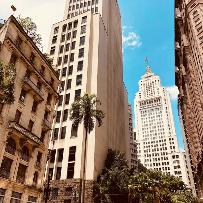 A cityscape featuring tall buildings and palm trees under a clear blue sky. The architecture includes both modern high-rises and historic structures. One prominent white skyscraper towers in the background.