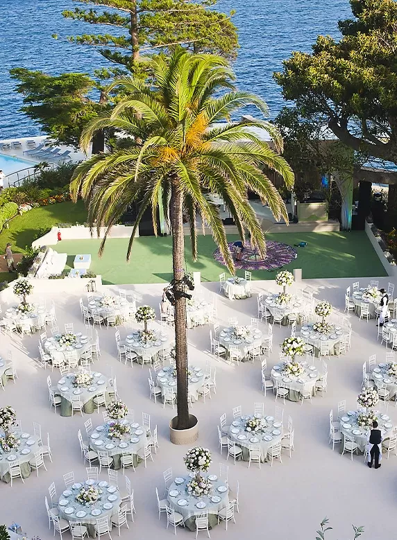 Outdoor wedding setup overlooking the ocean, featuring round tables with white tablecloths and floral centerpieces. A tall palm tree stands in the middle, surrounded by lush greenery and a clear blue sky.