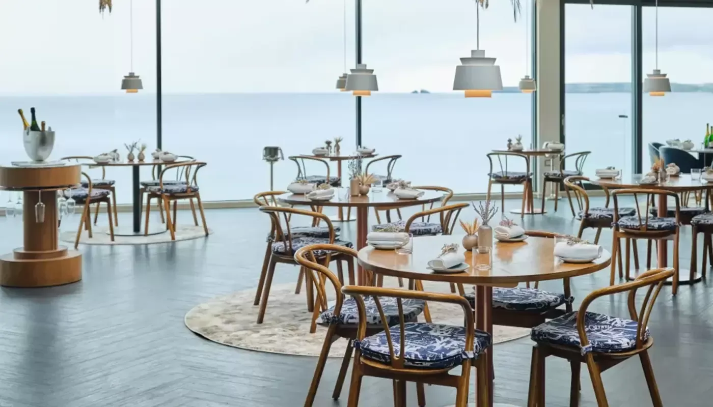 Adam Handling's michelin star luxury and sustainable restaurant. Open space with grey floor and wooden round tables with blue and white chair cushions sat on wooden chairs. The ceiling is white with hanging lights and a colorful display of flowers.