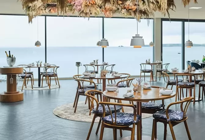 Adam Handling's michelin star luxury and sustainable restaurant. Open space with grey floor and wooden round tables with blue and white chair cushions sat on wooden chairs. The ceiling is white with hanging lights and a colorful display of flowers.