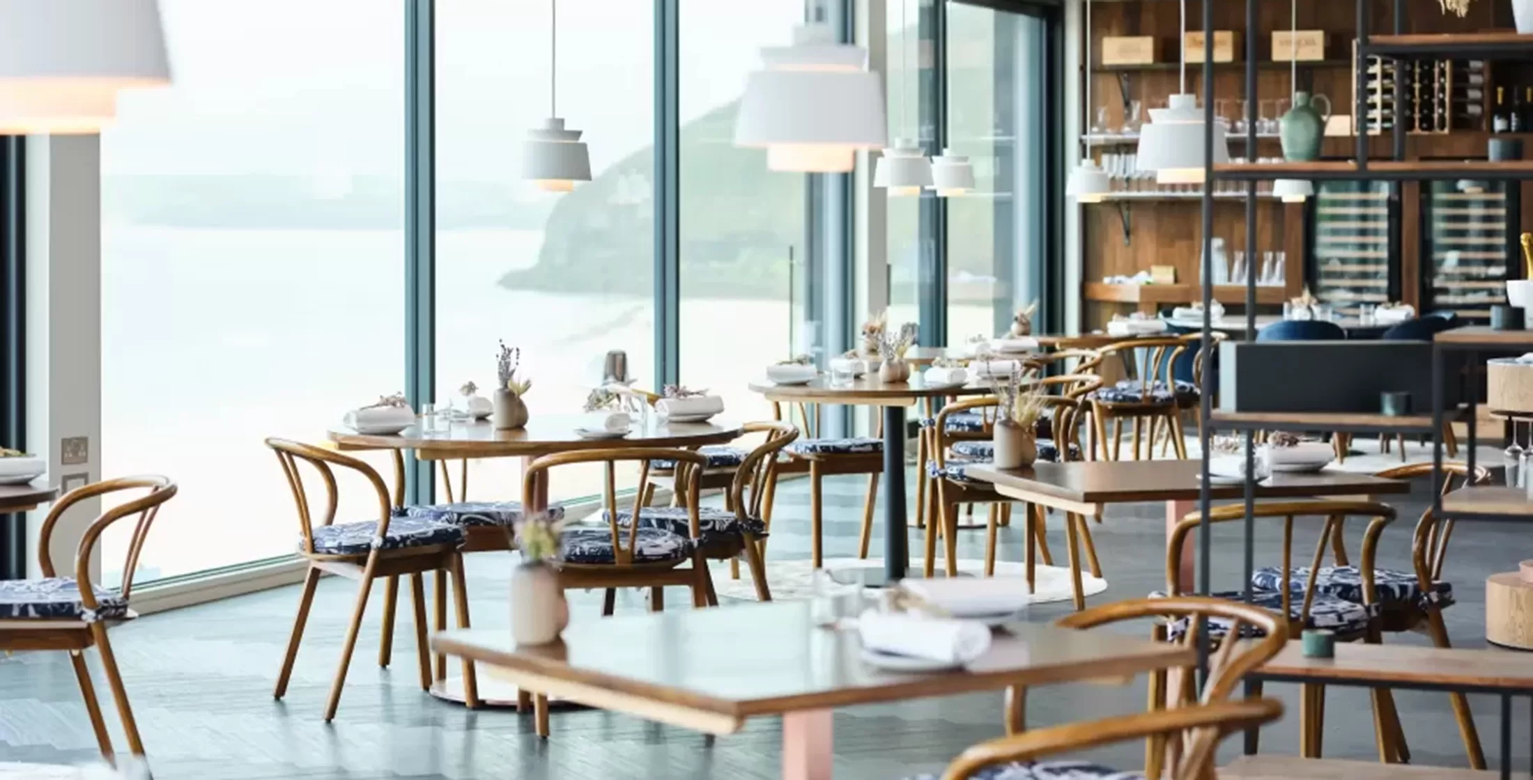 Adam Handling's michelin star luxury and sustainable restaurant. Open space with grey floor and wooden round tables with blue and white chair cushions sat on wooden chairs. The ceiling is white with hanging lights and a colorful display of flowers.