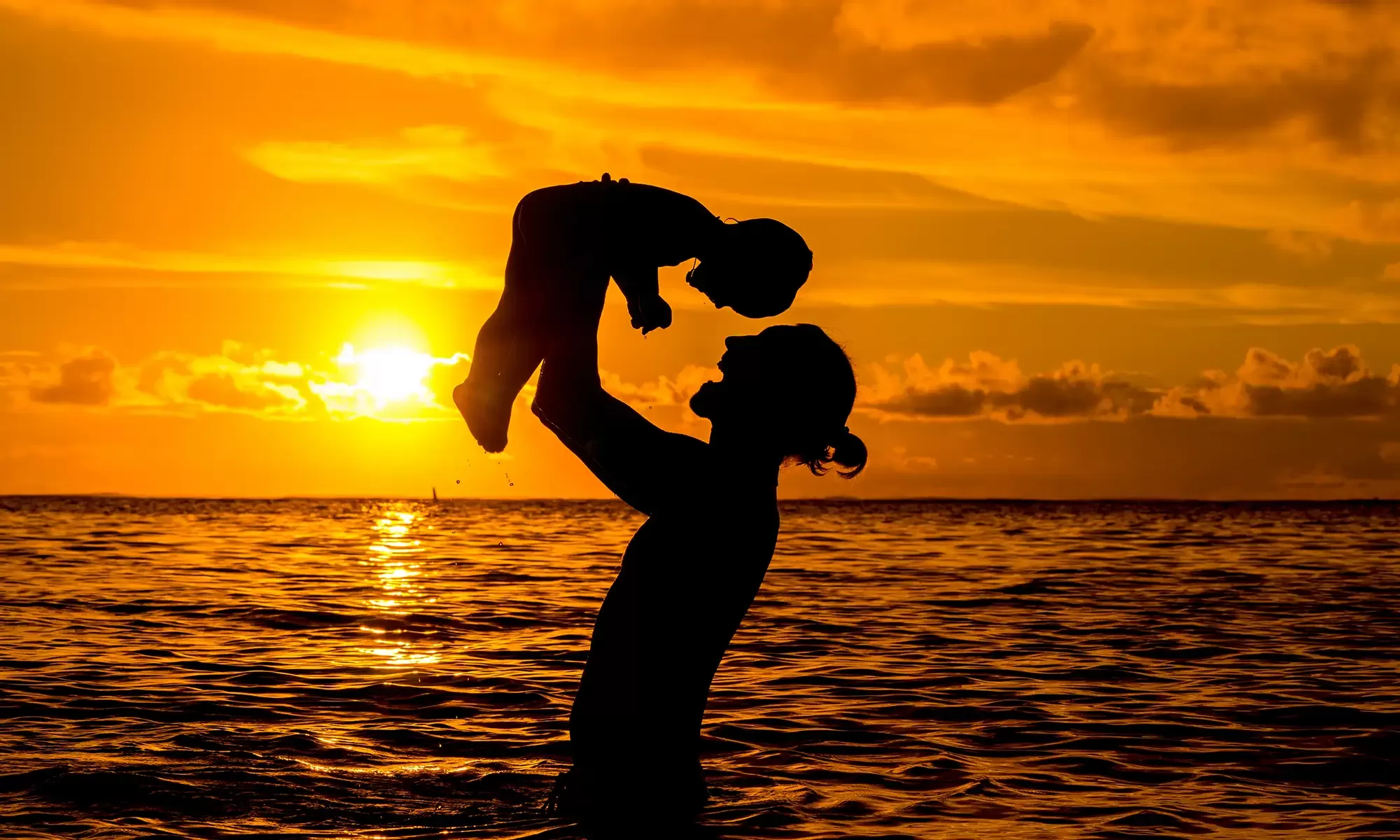 Silhouette of a person lifting a child against a vibrant sunset over the ocean. The sky is a mix of orange and yellow hues, with the sun near the horizon. The scene conveys warmth and joy, highlighting the bond between the two figures.