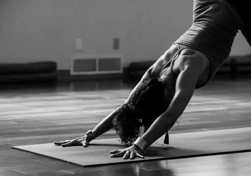A person performs the downward dog yoga pose on a mat in a dimly lit room. They are wearing a tank top and bracelets, with their head down and arms extended forward. The floor is wooden, and cushions are visible in the background.